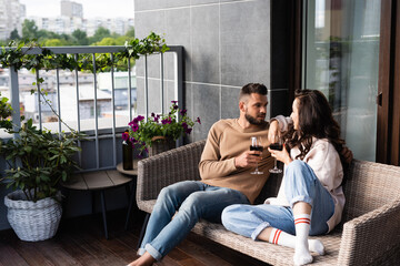 Obraz premium handsome man and attractive woman sitting on outdoor sofa and holding wine glasses while looking at each other