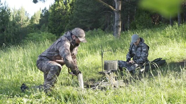 Man in camouflage clothes is chopping wood with axe and throwing it into bonfire. Tourist concept. Hunter checks or cleans hunting rifle. Best friends spend leisure weekend in forest nature background