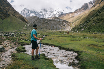 Fototapeta na wymiar Back view of male traveler dressed in active wear holding trekking sticks and admiring wildness nature of high mountains.Experienced tourist enjoying wanderlust standing near river in Salkantay
