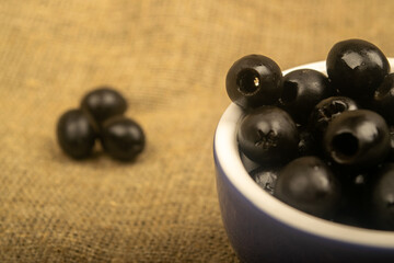 Black seedless olives in a ceramic bowl and a few scattered olives on a background of coarse-textured fabric. Close up.