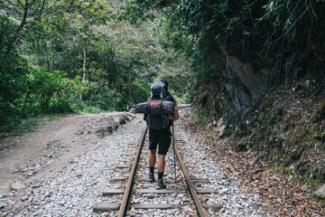 Obraz premium Rear view of tourist with backpack walking on railroad in mountains during wanderlust in wildness nature.Hiker with trekking sticks exploring environment of south landscapes during hiking trip