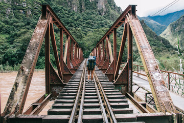 Fototapeta na wymiar Back view of young woman tourist with backpack discover new places in wildness environment crossing bridge with rails over river.Hiker with travel rucksack going in mountains during trek