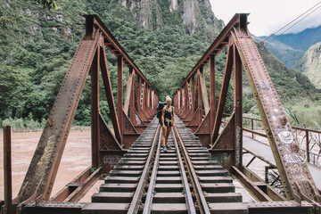 Fototapeta na wymiar Portrait of positive young woman with tourist backpack smiling at camera and standing on rails of bridge while travelling in wildness nature with high mountains and beautiful scenery on background