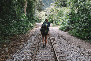 Obraz premium Back view of professional female hiker with tourist backpack exploring wilderness environment during hiking trekking on railway in destination of mountains and green vegetation to discover new places