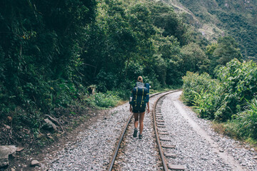 Fototapeta premium Rear view of female tourist with backpack discovering new places in wildness environment of mountains.Hiker with rucksack going on rainway and exploring green vegetation during trekking holiday