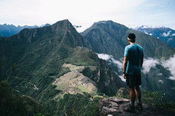 Naklejka premium Back view of young man dressed in active wear climbed on peak of mountain and admiring beautiful scenery of nature.Experienced male tourist enjoying amazing green hills during trip