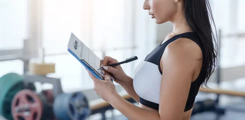 Tuinposter Personal trainer filling out workout planner at sports club, copy space © Prostock-studio