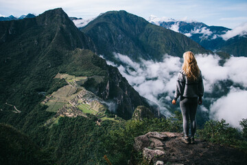 Naklejka premium Back view of young woman tourist standing on high peak of mountain and enjoying amazing scenery of hills and white clouds during trekking trip on weekend.Female hiker admiring beautiful nature