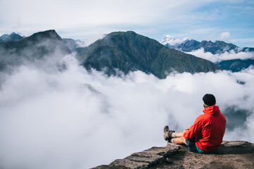 Rear view of male tourist dressed in active wear sitting on rocky and enjoying amazing scenery of environment and travel lifestyle during trek.Hiker relaxing and admiring beauty of green mountains