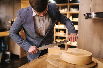 Man slicing a cheese on wooden board. Cheese shop. Man use big cheese knife