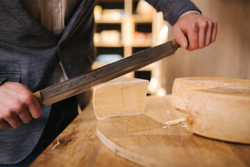 Man slicing a cheese on wooden board. Cheese shop. Man use big cheese knife - obrazy, fototapety, plakaty