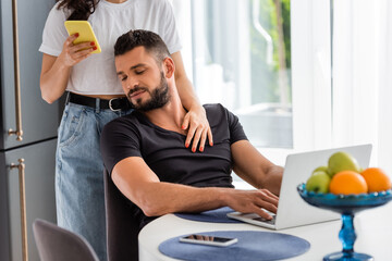 selective focus of woman standing and touching bearded freelancer boyfriend near laptop while using smartphone