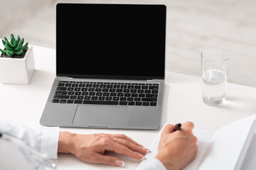 Woman doctor hands making notes in notebook, using laptop, sitting at desk