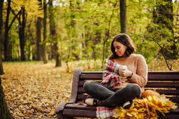 A girl holds a coffee and sits on a bench in the park in the fall. on the bench beautiful autumn...