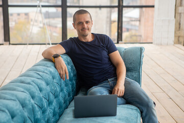 Man with laptop sitting on sofa, looking in camera and smiling.