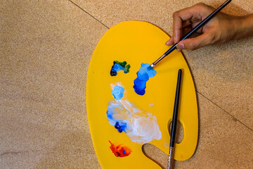 Oil painting palette full of colors and brushes kept on floor . Hand with a brush is mixing colors