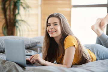 Happy woman using laptop computer while lying on bed in morning