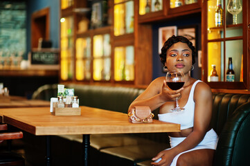 African american woman, retro hairstyle in white dress at restaurant with glass of wine.