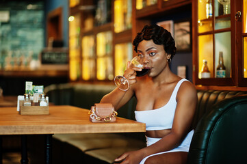 African american woman, retro hairstyle in white dress at restaurant with glass of wine.