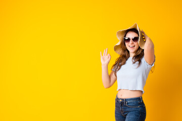 Obraz na płótnie Canvas Happy smiling brunette girl showing ok okay gesture and holding her summer straw hat,wearing blue t-shirt and jeans,sunglasses isolated over yellow background.Approval sign.Body language.Copy space