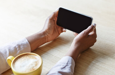 Cropped view of young woman holding smartphone with blank screen while drinking coffee at cafe, mockup for design