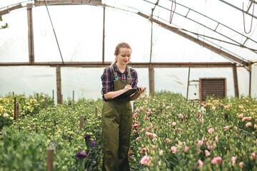 Fototapeta na wymiar Gardener woman working on flowers in greenhouse and makes notes. Floral business concept.