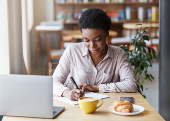 Fototapeta na wymiar African American female student making notes in copybook while studying online at cafe