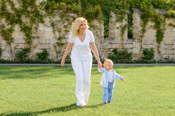 Fototapeta premium Mom leads her son's hand through the green grass. Both blondes.