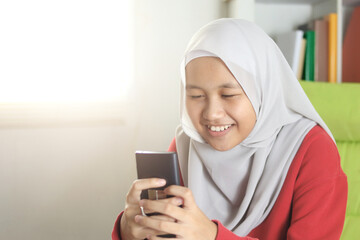 Happy Asian muslim girl wearing hijab smiling while browsing trough internet or reading chat message on her phone