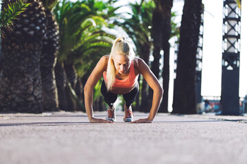 Fototapeta premium Young caucasian woman doing press ups in tropic urban setting with palm trees, athletic female runner with beautiful figure dressed in workout gear doing push-ups outdoors before begin her morning run