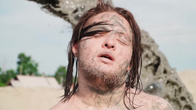 Portrait Funny Fat Man with Long Hair Bathes in the Mud. Against the background is pouring Black Dirty Water from the Pipe. Close up. Slow motion