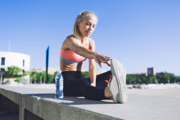 Fototapeta na wymiar Young attractive fit woman with beautiful figure stretching legs while working out in urban setting at sunny day, caucasian female jogger doing warming up exercise before began her morning run