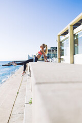 Young female runner dressed in tracksuit enjoy rest and sun after intense exercising outdoors while sitting on pier against blue sky background with copy space area for text message or advertising