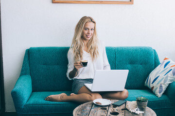 Successful hipster girl sitting on couch in living room using laptop device and wireless connection...