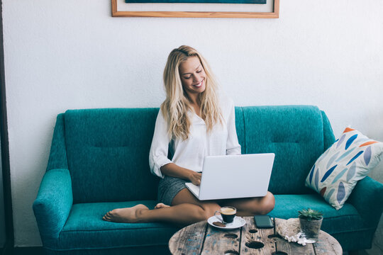 Cheerful woman sitting on cozy sofa and tying email via modern laptop device, barefoot smiling hipster girl enjoying messaging with friends in cozy home interior while share publication in blog