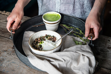 Woman holding metal tray with natural yogurt with granola and different seeds in white ceramic bowl, spoon, matcha latte  and eucalyptus branch.