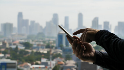 A man dials a message on a smartphone against the background of the panorama of the city of Jakarta. Hands close up.