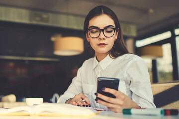 Fototapeta na wymiar Confident attractive female owner of trading corporation making paperwork and checking email waiting for message from marketing experts providing advertising campaign on popular website via smartphone