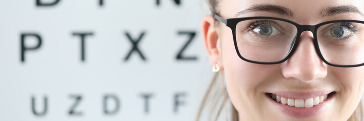 Close-up of cheerful female ophthalmologist smiling on camera. Special board with letters to checkup vision. Copy space in left side. Modern medicine healthcare and eye doctor concept - obrazy, fototapety, plakaty