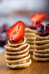 Stacks of mini pancakes with strawberry and cherry topping are laying on wooden cutting board at kitchen