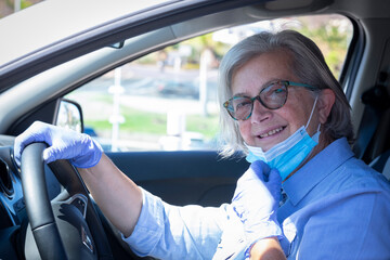 Senior woman with sunglasses  driving her silver car with facial mask and protective gloves due to coronavirus - new normal life until the end of the pandemic