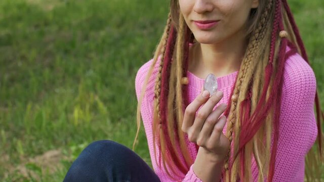 Adult girl in knitted sweater with long dreadlocks is holding transparent violet amethyst yoni egg for vumfit, imbuilding or meditation outdoors on her body background outdoors