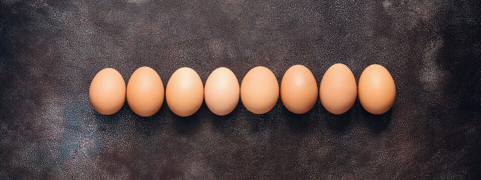 Brown chicken eggs in a row on a dark painted rustic background. Top view, flat lay, copy space, border.