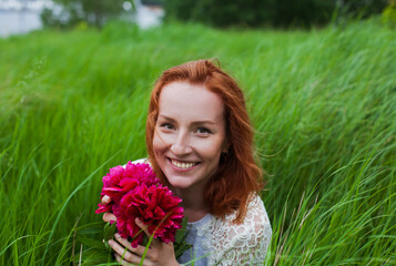 Fototapeta premium Smiling redhead girl with peonies in green grass