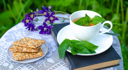 A Cup of green tea with a mint leaf, flowers, and a book on a table in the garden. Evening tea with cookies. Summer village concept. Vintage banner.
