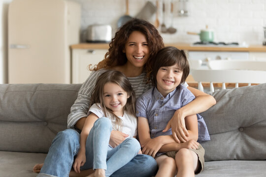 Close up portrait picture of happy young mother with children sitting on couch at home. Smiling parents with little kids hugging looking at camera posing for photo in living room.