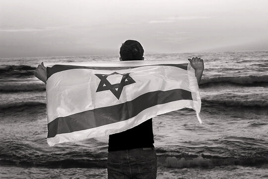 Israeli patriot with the flag of Israel stands on the shores of the Sea at sunset. Independence day Israel, patriotic holiday concept. Dramatic monochrome black and white photography