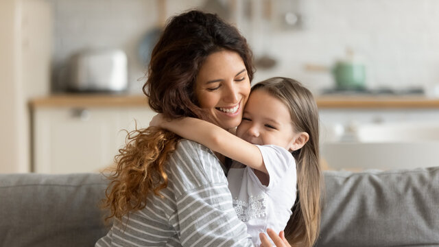 Close up happy attractive mother hugging cute preschool daughter. Smiling young mom closed eyes embracing little girl enjoying free time together sitting on couch at home.