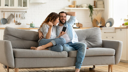 Obraz premium Smiling man and woman hugging using smartphone reading joke and laughing sitting on couch in living room. Loving husband embracing young wife holding smartphone in hand watching funny video at home.