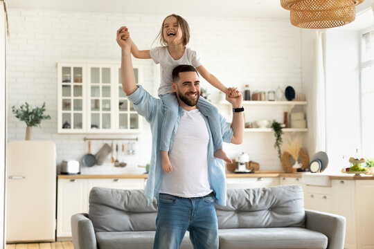 Happy little daughter sitting on father neck piggybacking playing active game at home. Smiling dad holding cute girl with arms outstretched carrying on back. Family spending weekend together.